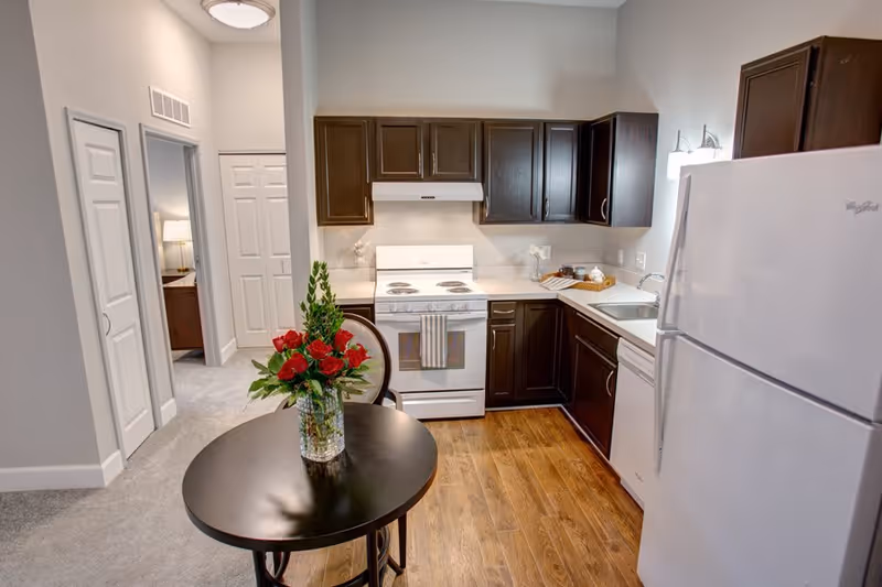 A modern kitchen area with dark wood cabinets, a white electric stove, a white refrigerator, and a white dishwasher. There is a small round black table with a vase of red roses and greenery in the foreground. The floor is wooden, and the walls are light-colored. A doorway leads to another room with carpeted flooring and a lamp visible.
