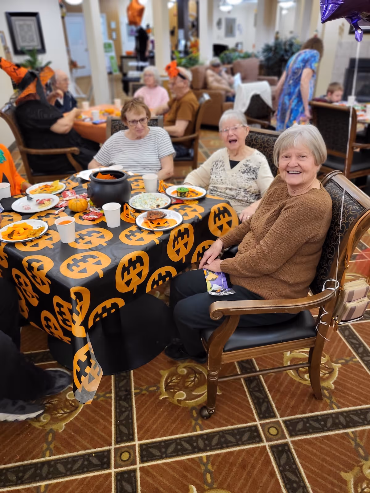 A group of elderly people sitting around a table decorated with a black tablecloth featuring orange jack-o'-lantern faces. The table has plates of food and paper cups, and some people are wearing Halloween-themed accessories. The setting appears to be a communal dining or activity area in a senior living facility.