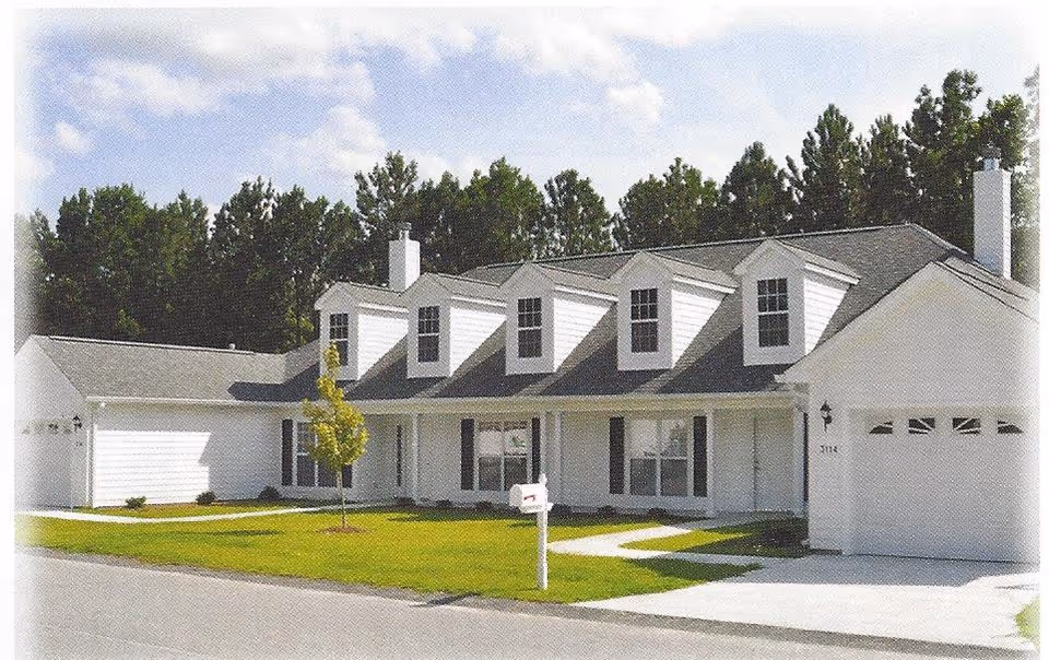 Exterior view of a white residential building with multiple dormer windows on the roof, a green lawn, a small tree, and a white mailbox in front. The building has a garage on the right side and is surrounded by tall trees in the background under a partly cloudy sky.