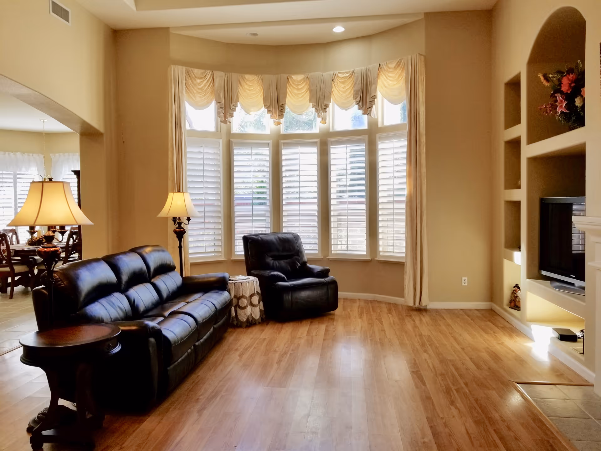 A cozy living room with a black leather sofa and matching armchair, a round side table with a patterned cloth, two standing lamps with beige lampshades, large windows with white plantation shutters and beige curtains, built-in shelves with decorative items, and a flat-screen TV. The room has light wood flooring and beige walls.