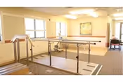 A rehabilitation or physical therapy room with parallel bars for walking exercises, beige walls, carpeted floor, windows letting in natural light, and a framed abstract painting on the wall.