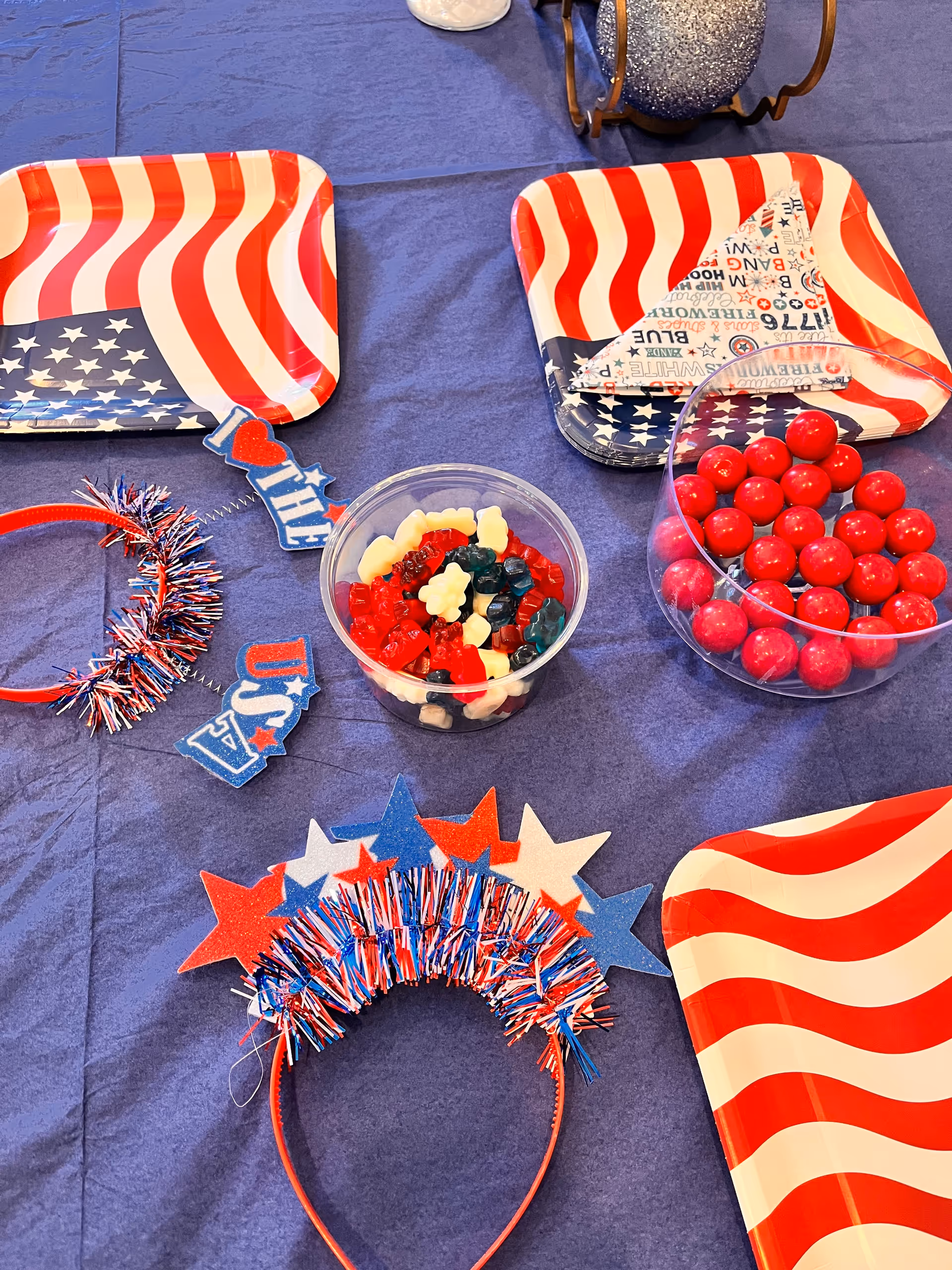 A table set with patriotic-themed items including red, white, and blue star-shaped headbands, decorative signs with USA and I love the USA messages, bowls of red gumballs and gummy bears, and plates with American flag designs on a blue tablecloth.