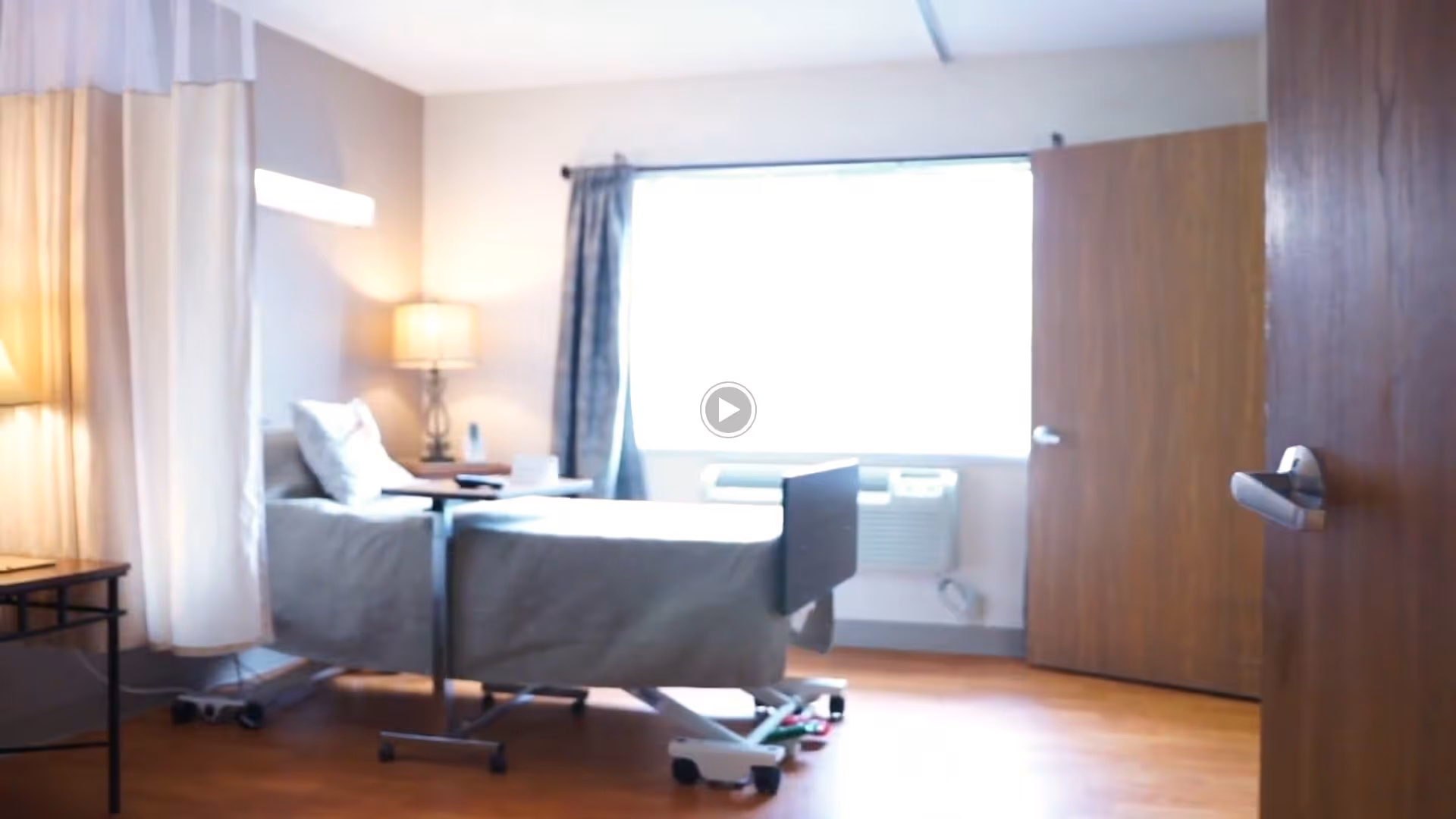 A bright room with a hospital-style bed, a bedside table with a lamp, a curtain partially drawn on one side, a large window with curtains, and a wooden door slightly open.