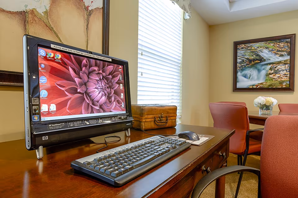 A desktop computer with a large monitor displaying a red flower wallpaper sits on a wooden desk with a keyboard and mouse. Behind the desk, there is a window with blinds, a wooden box, and a table with red chairs and a vase of white flowers. A framed picture of a flowing stream hangs on the wall.