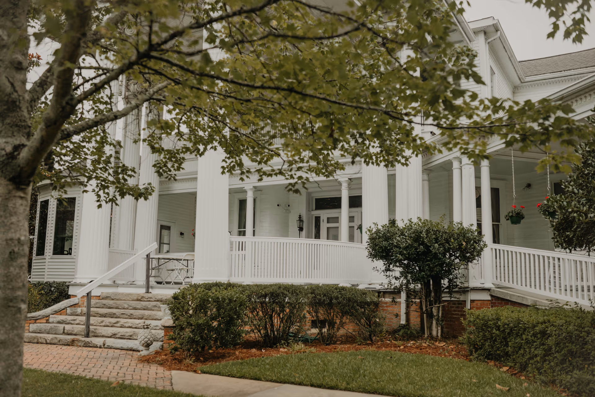 Front exterior view of a large white building with tall columns, a porch with a ramp and stairs, surrounded by green bushes and trees.
