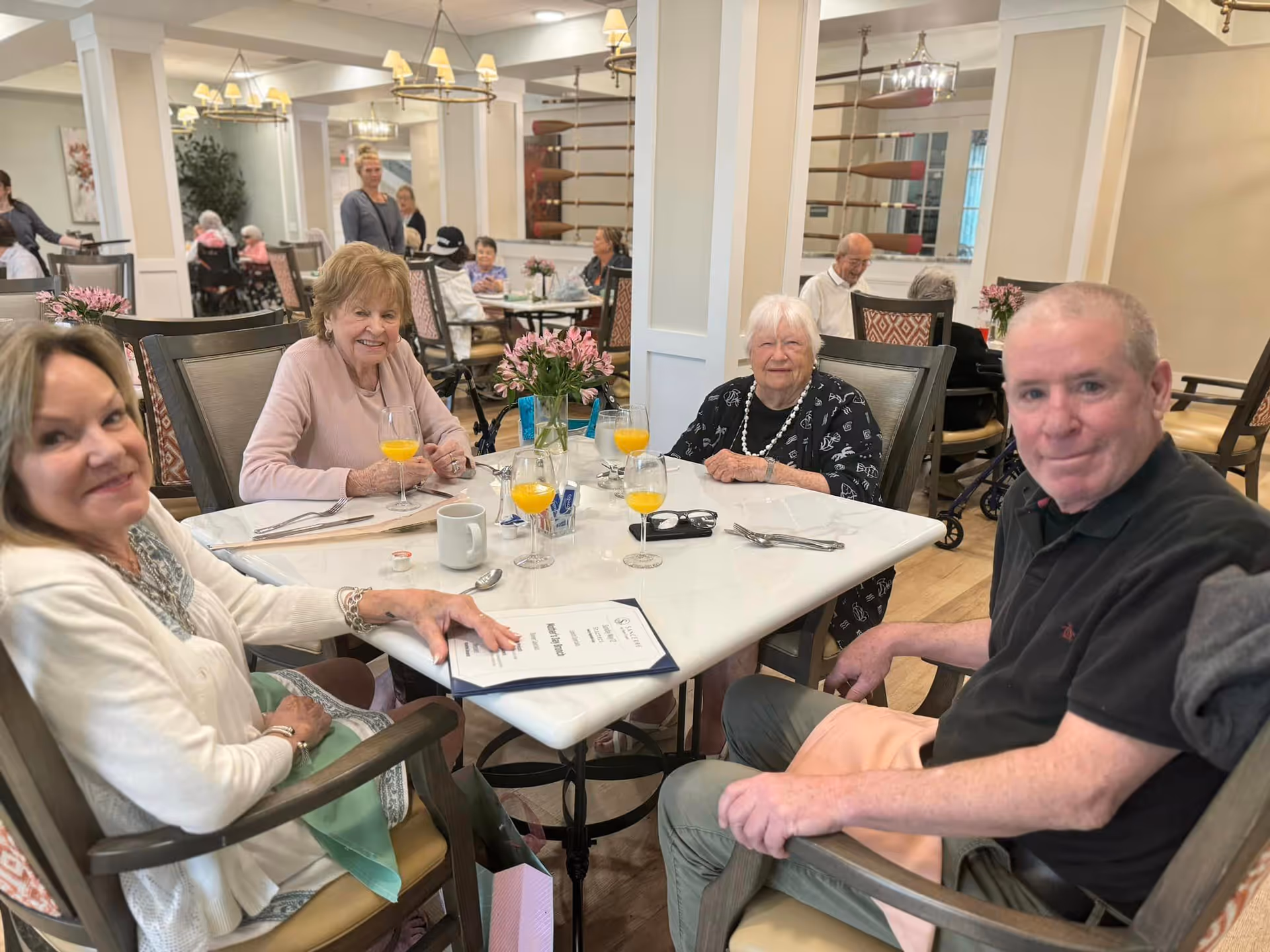 Several senior residents sit around a table with glasses of orange drink in a bright communal dining room.