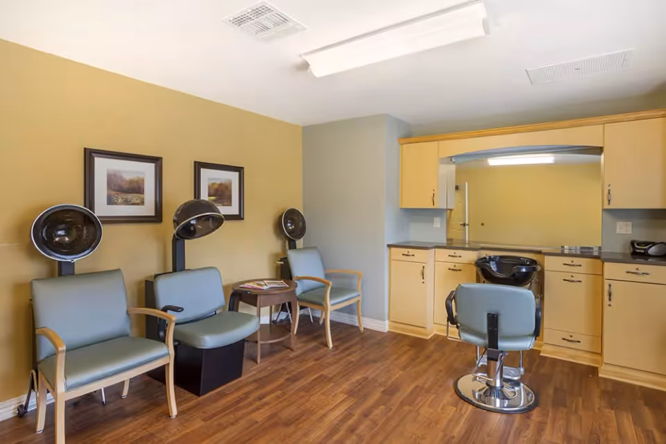Interior view of a hair salon area in an assisted living facility with three hair drying chairs along a wall, a small table with magazines, a salon chair in front of a large mirror, and wooden cabinets underneath the mirror. The room has wood flooring and light-colored walls with two framed pictures.