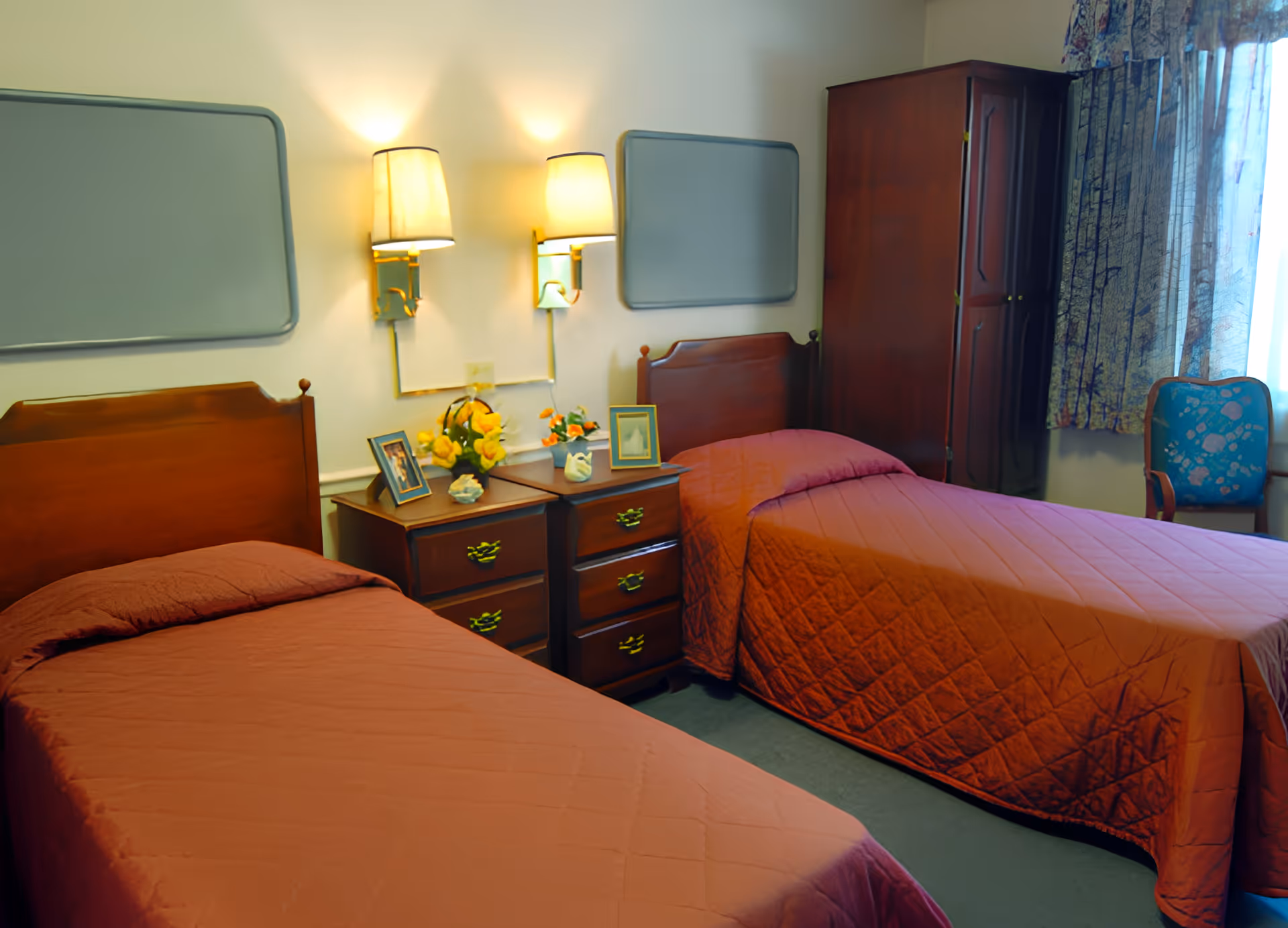 A bedroom with two single beds covered in reddish-orange quilts. Between the beds is a wooden nightstand with drawers, decorated with framed photos and flower arrangements. Above the nightstand are two wall-mounted lamps and two rectangular mirrors. To the right, there is a wooden wardrobe and a window with patterned curtains and a blue chair beside it.
