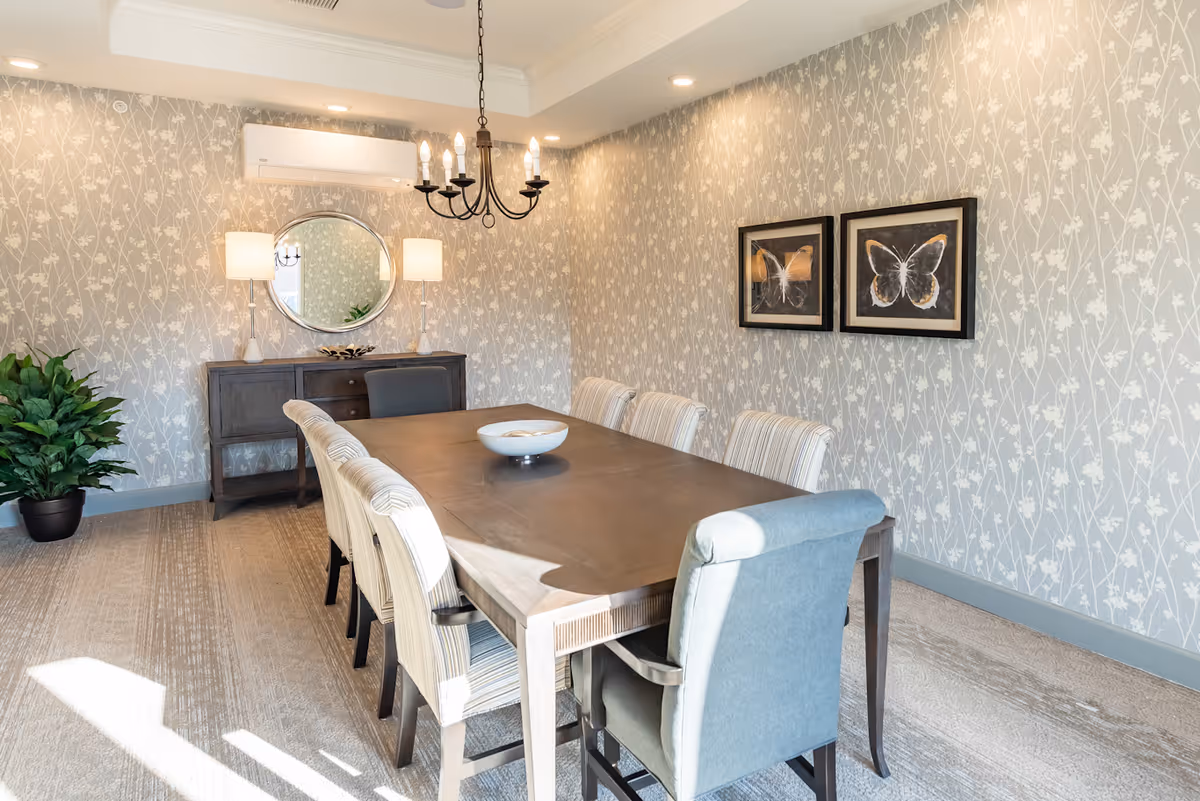 A bright dining room with a long wooden table surrounded by upholstered chairs, a sideboard with mirror and lamps, chandelier, and butterfly artwork.