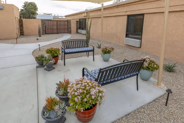 Outdoor patio area with two black metal benches with blue patterned cushions, surrounded by several potted plants and flowers. The patio is adjacent to a beige stucco building with windows and air conditioning units. The ground is covered with gravel and a concrete walkway curves through the area.