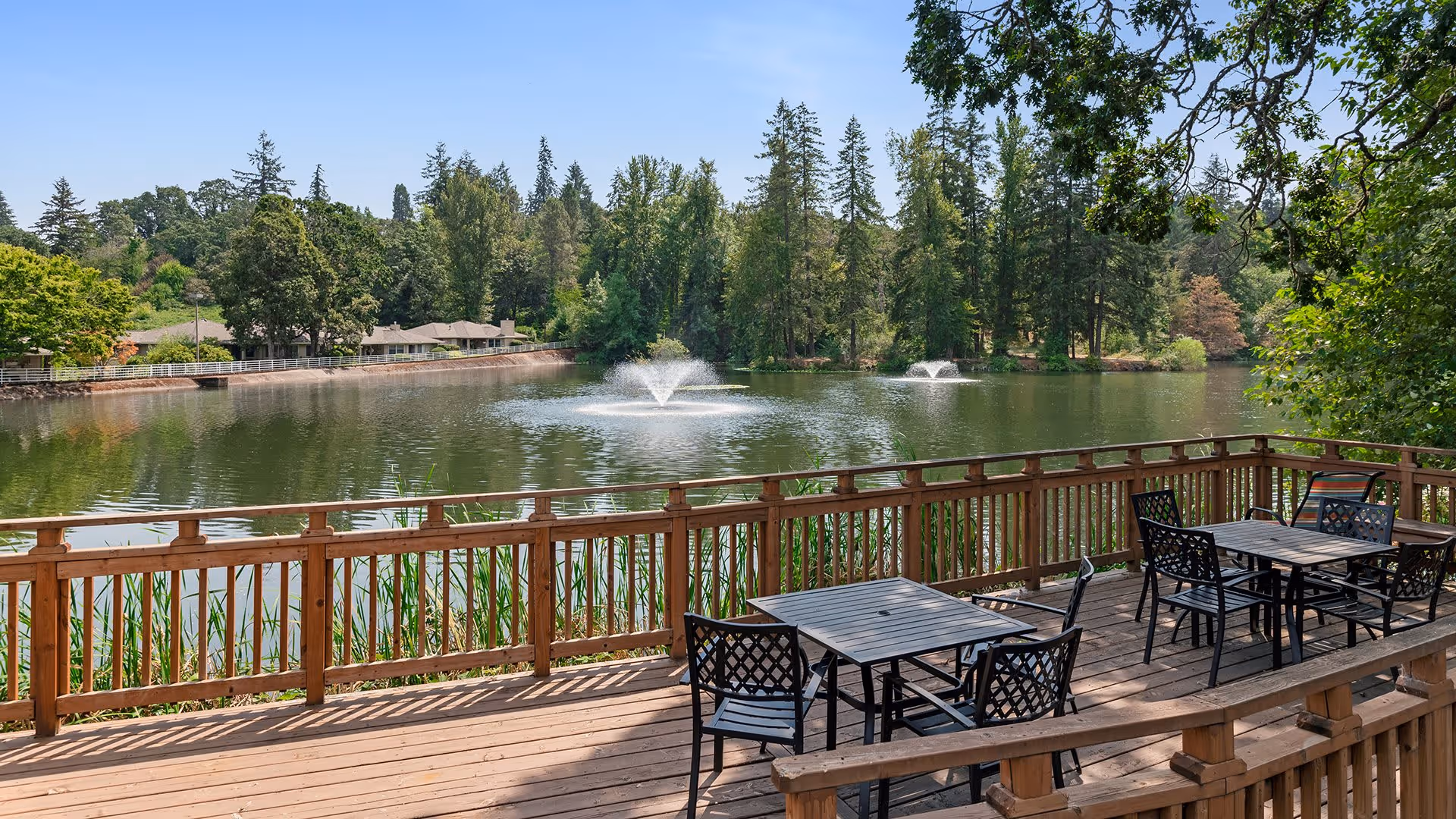 Outdoor wooden deck with several black metal tables and chairs overlooking a lake with two water fountains, surrounded by trees and greenery under a clear blue sky.