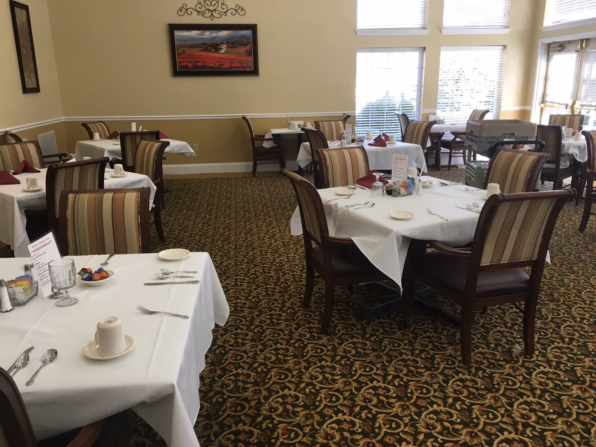 Dining room with white-clothed tables and striped upholstered chairs arranged on a patterned carpet near large windows.