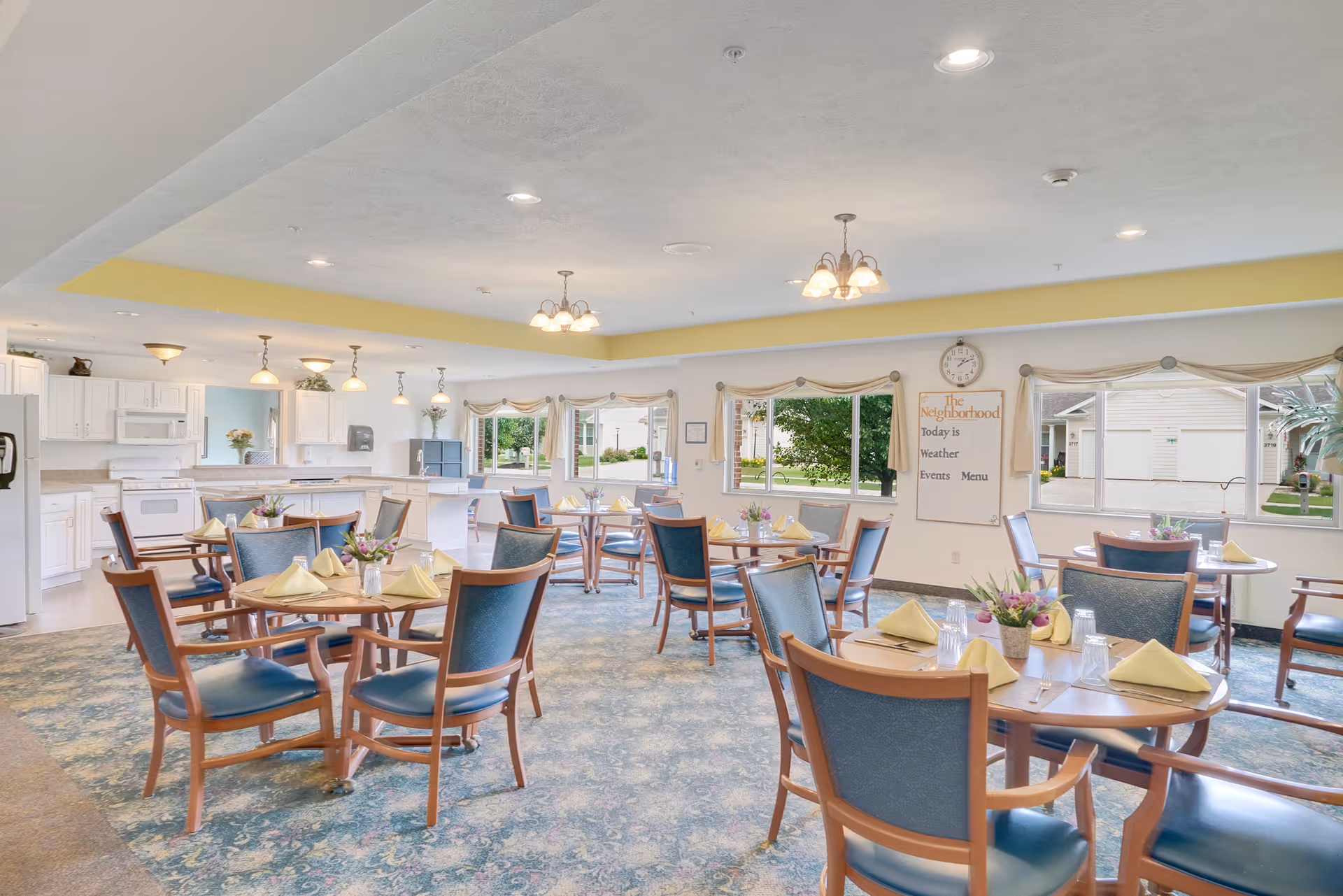 Bright communal dining room with round tables set with napkins and centerpieces and a kitchen area in the background.