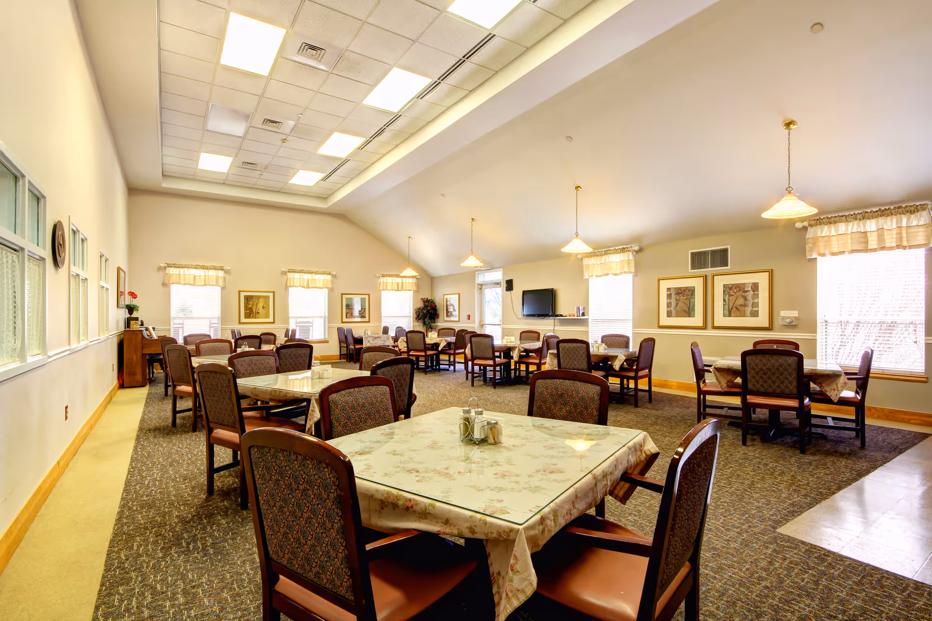 A spacious dining room with multiple tables covered with floral tablecloths and surrounded by cushioned chairs. The room has large windows with valances, framed artwork on the walls, hanging pendant lights, a television mounted on the far wall, and a piano in the corner. The ceiling is high with recessed lighting.