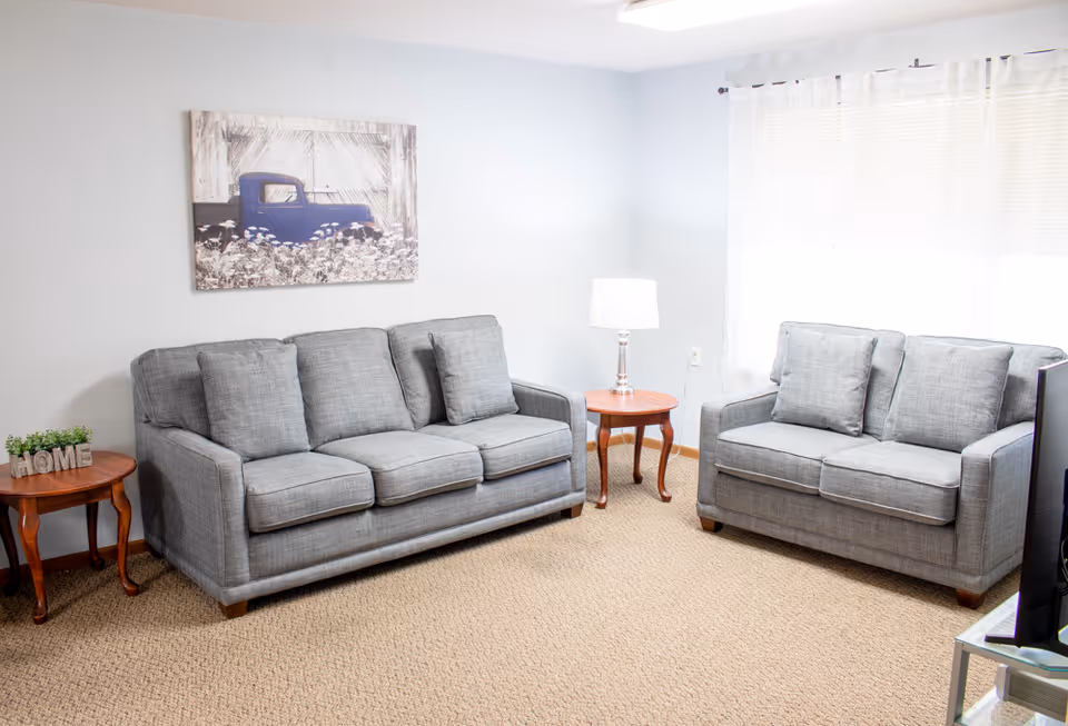 Bright living room with two gray sofas, wooden side tables, a lamp, wall art, and a window with blinds.