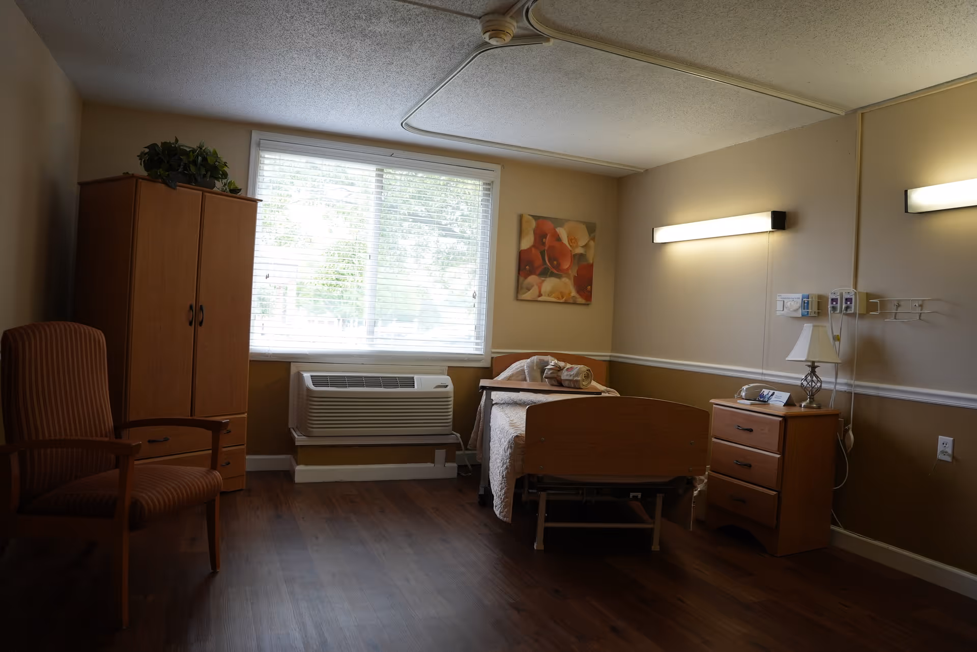 A simple, clean senior living bedroom with a single bed, wooden nightstand with a lamp and phone, a wooden wardrobe, a striped armchair, and a large window with blinds letting in natural light. The walls are beige with a floral painting and two wall-mounted lights.