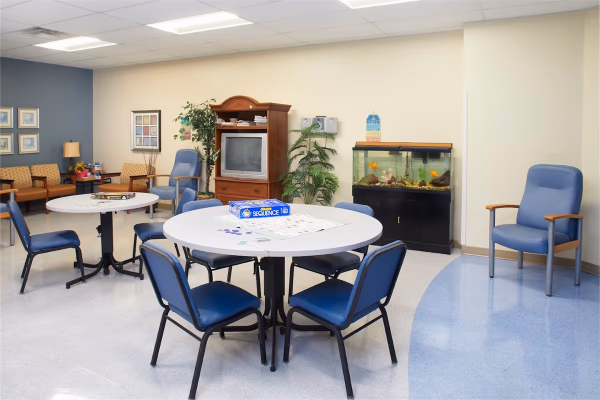 A senior facility common room with round tables and chairs, a TV cabinet, aquarium, and additional seating.