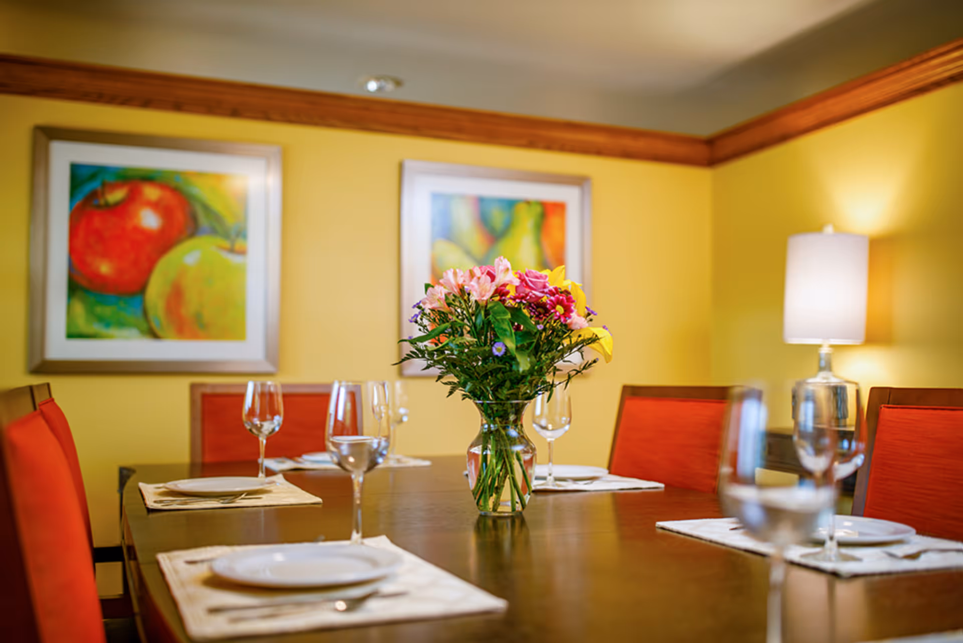 A dining room with a wooden table set with white plates, wine glasses, and silverware on beige placemats. A vase with a colorful bouquet of flowers is placed in the center of the table. The room has yellow walls with two framed paintings of fruit and a table lamp with a white shade in the background. The chairs have red cushions.