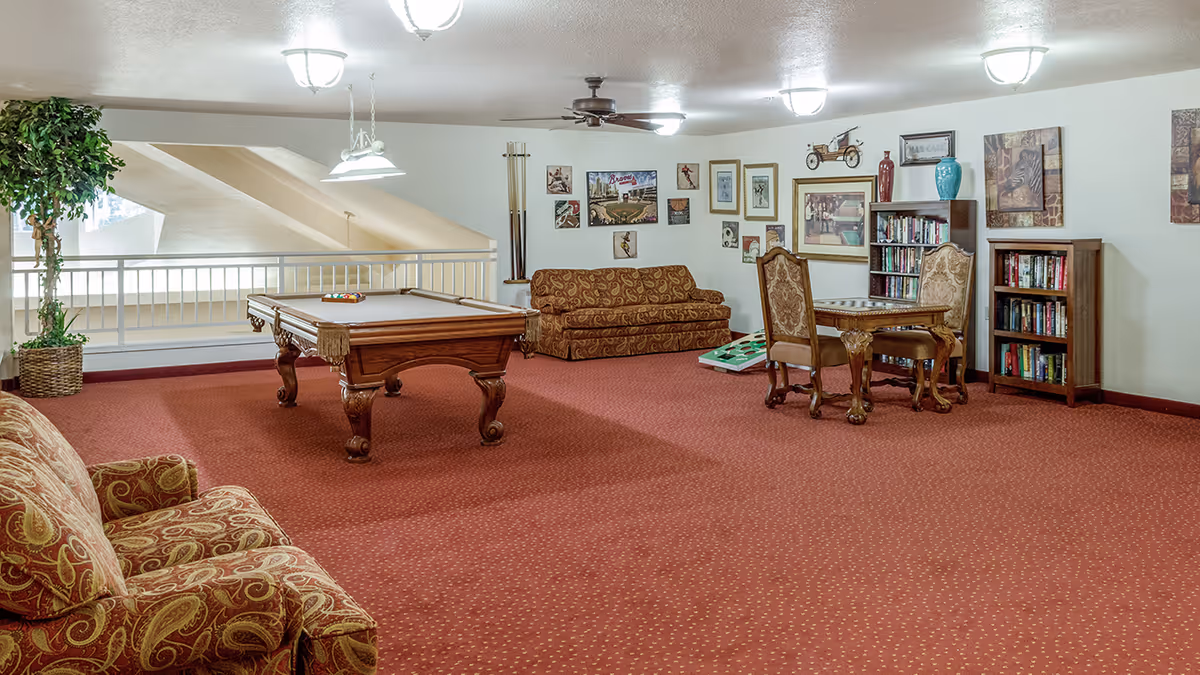 A spacious common area with red carpet featuring a pool table, two patterned sofas, a small table with two ornate chairs, bookshelves filled with books, and various framed pictures on the walls. There is a ceiling fan and multiple ceiling lights illuminating the room.