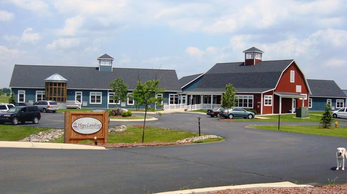 Exterior view of Hope Landing assisted living facility showing a large building with a red barn-style section and a blue section, surrounded by a paved driveway and parking area with several cars parked. There is a wooden sign in front displaying the facility name, some small trees, and a dog standing on the right side of the driveway.