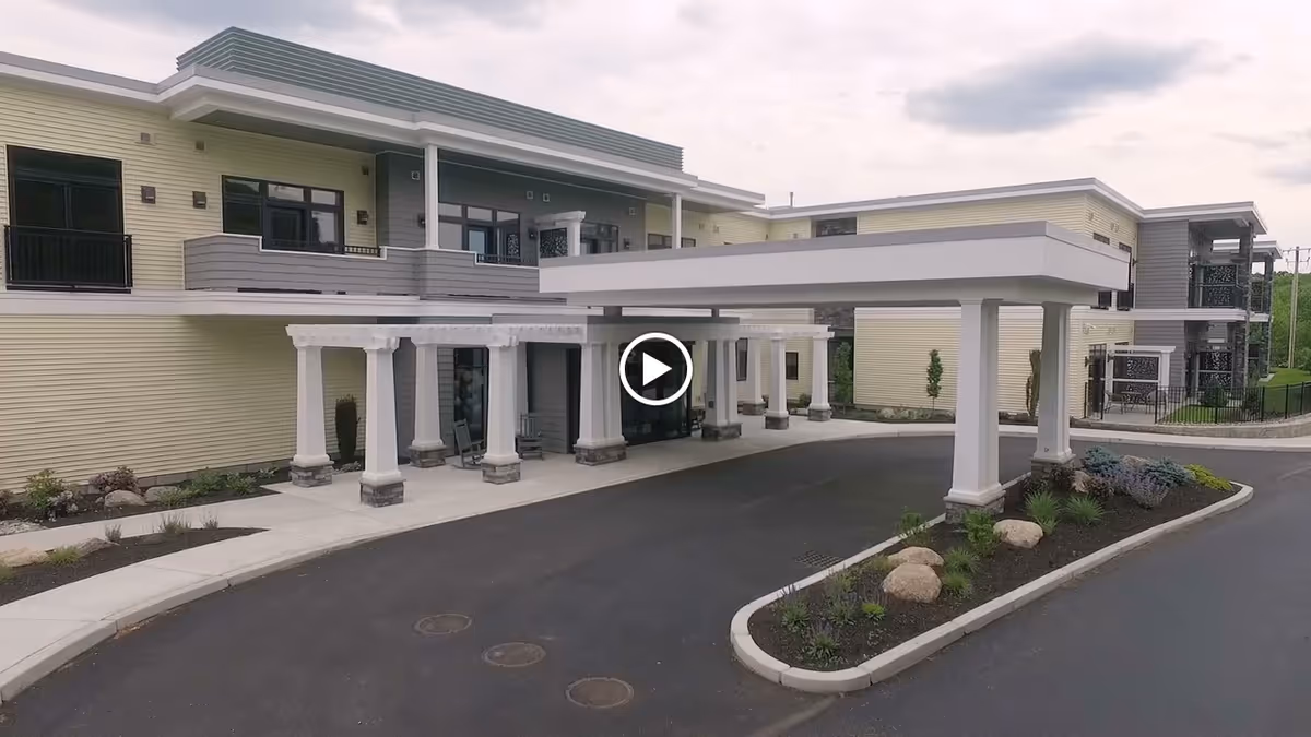 Front entrance of a two-story assisted living building with a covered porte-cochere and landscaped driveway.