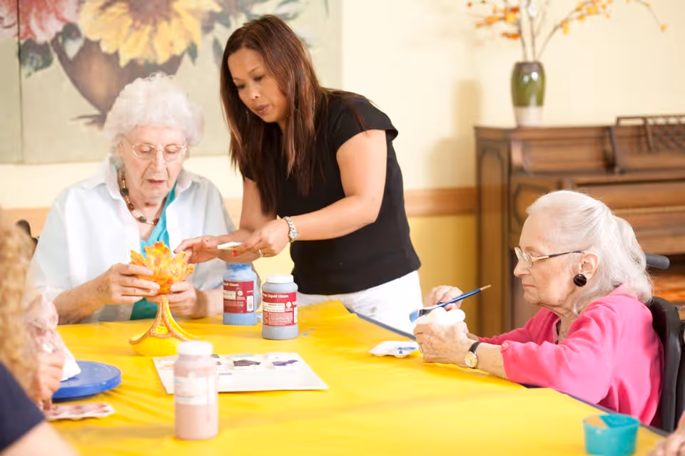 An elderly woman and a caregiver are engaged in a painting activity at a table covered with a yellow tablecloth. The elderly woman is holding a painted ceramic piece shaped like a tree or flower, while the caregiver is assisting her. Another elderly woman in a pink sweater is painting a small object on the right side of the table. There are paint bottles and brushes on the table, and a piano and a vase with flowers are visible in the background.
