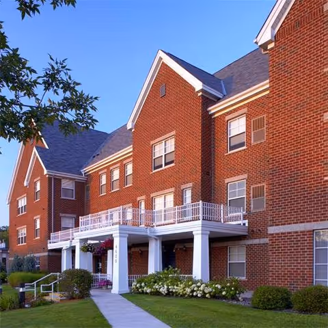 Exterior view of a red brick senior living facility building with white trim and a covered entrance supported by white columns. The building has multiple windows, a balcony with white railings, and a well-maintained lawn with bushes and flowers along a concrete walkway leading to the entrance. The sky is clear and blue.