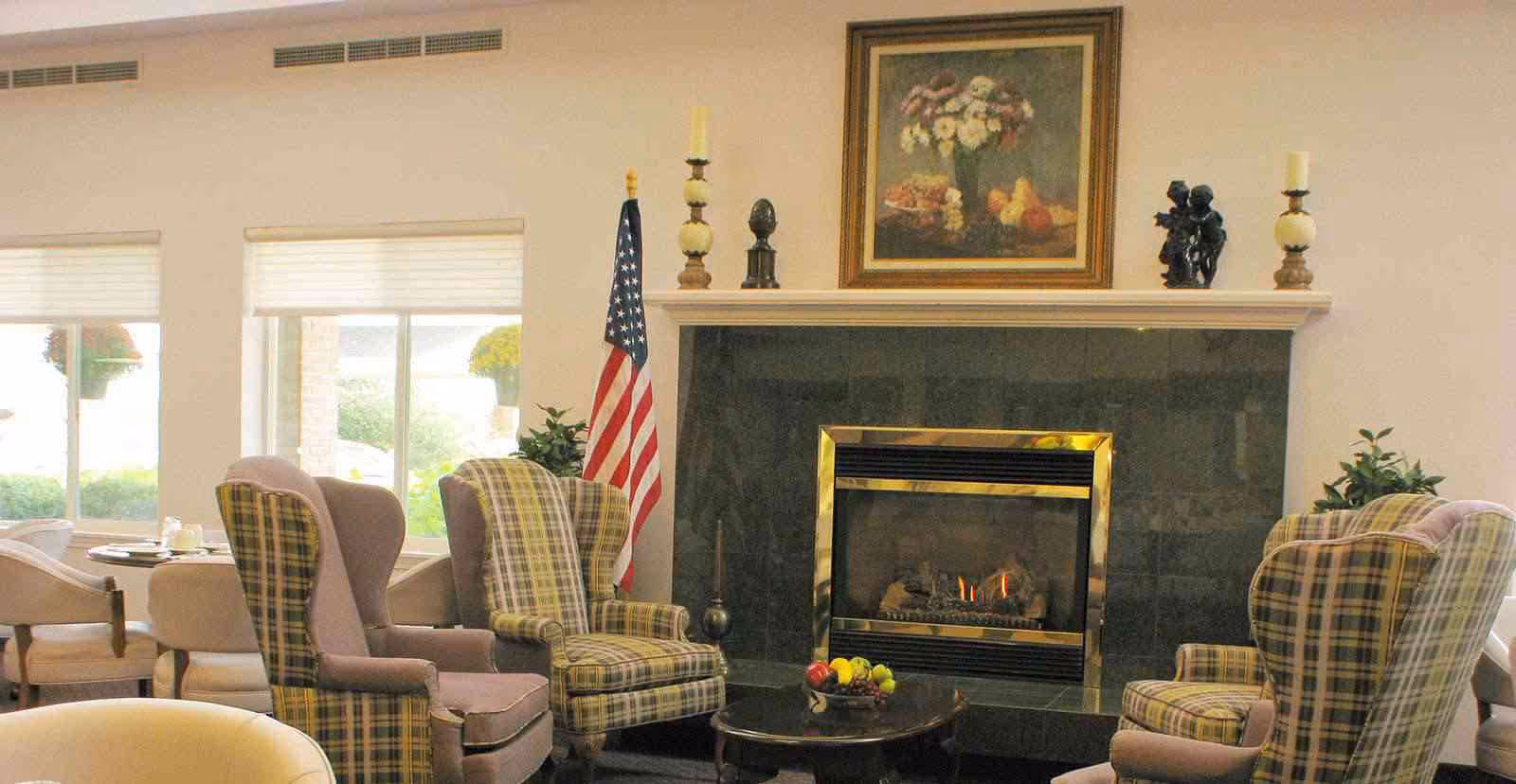 A cozy senior living common area with a fireplace surrounded by four plaid armchairs. On the mantel above the fireplace are decorative items including candles, a small bust, and a sculpture, with a framed painting of flowers hanging on the wall. An American flag stands to the left of the fireplace, and windows with white blinds allow natural light into the room. A round coffee table with a bowl of fruit is in front of the fireplace.