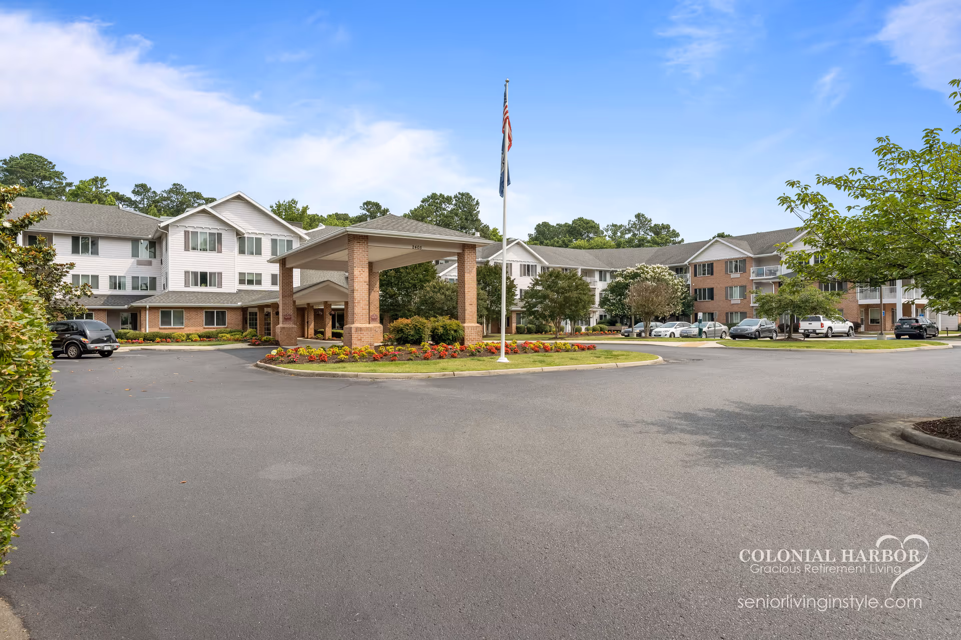 Exterior view of Colonial Harbor Gracious Retirement Living facility showing a large three-story building with a covered entrance, surrounded by landscaped flower beds and trees, with several parked cars and a flagpole with the American flag.
