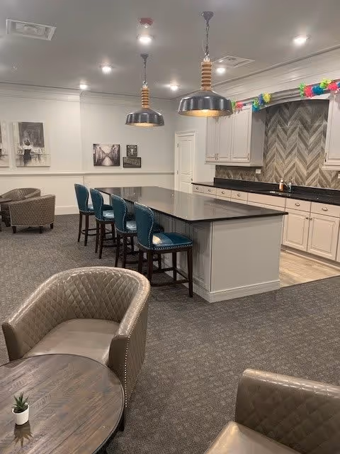 Interior view of a senior living facility common area featuring a kitchen island with four blue cushioned bar stools, two large pendant lights hanging above, beige cabinetry, and a herringbone patterned backsplash. In the foreground, there are two quilted leather armchairs and a small round wooden table with a small plant on it. The room has carpeted flooring and neutral-colored walls with framed artwork.