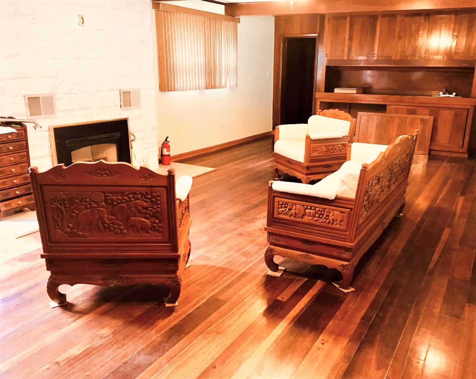 Wood-paneled living room with carved wooden sofas on polished hardwood floors, a fireplace and a small built-in bar.