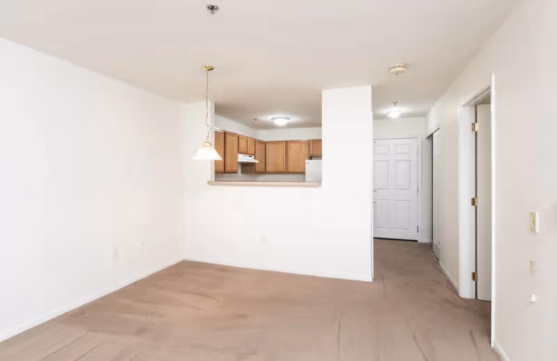 Empty apartment interior showing a small kitchen with wooden cabinets and white appliances, a hanging light fixture, beige carpeted floor, white walls, and a white front door in the background.