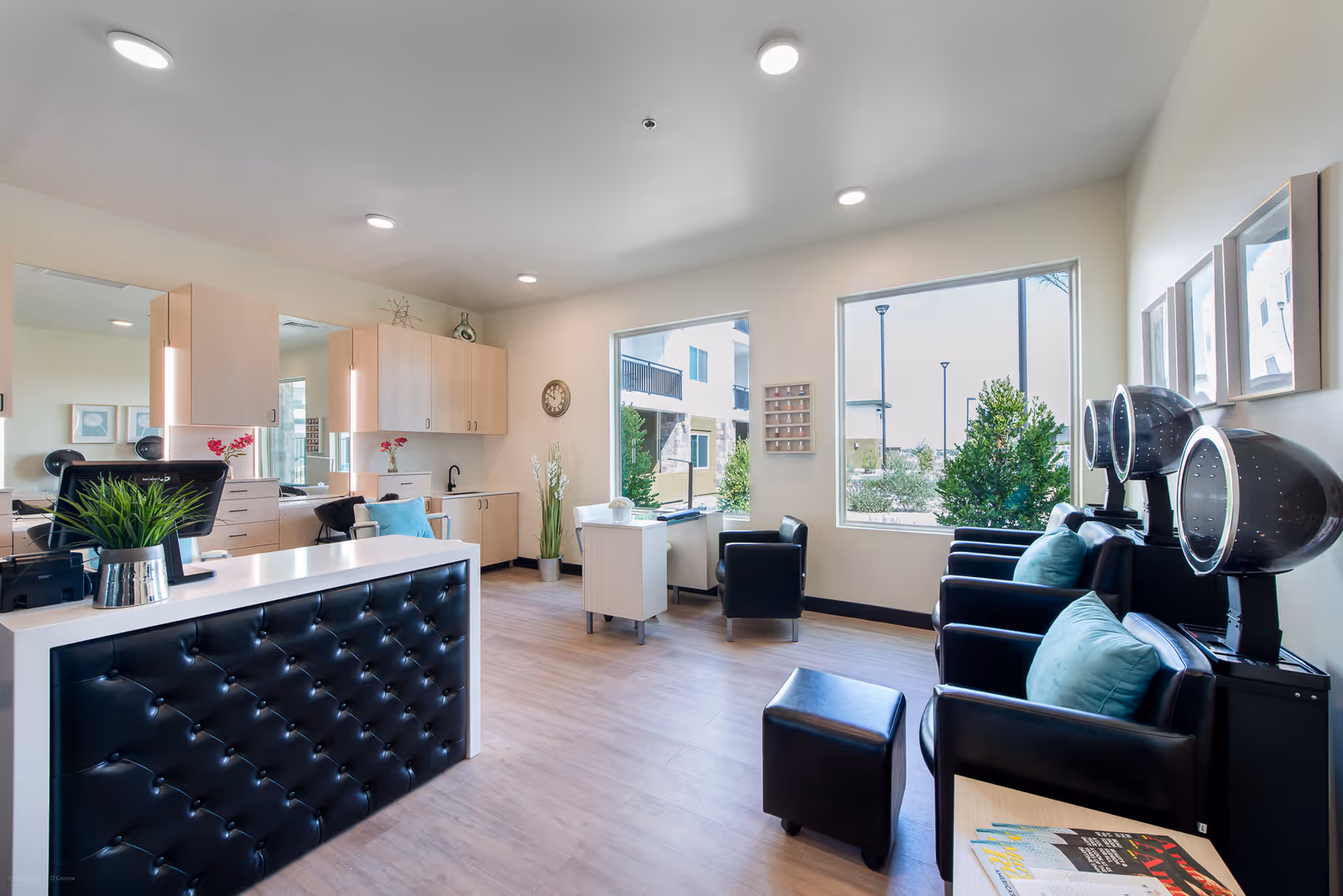A bright and modern salon area with black leather chairs equipped with hair dryers, a reception desk with a tufted black front panel, light wood cabinetry, large windows showing outdoor greenery, and a small table with magazines.