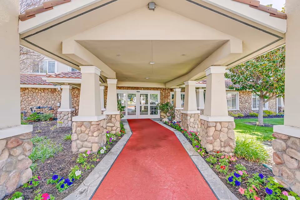 Covered entrance walkway with stone pillars and a red carpet leading to double glass doors of a building, surrounded by flower beds and greenery.