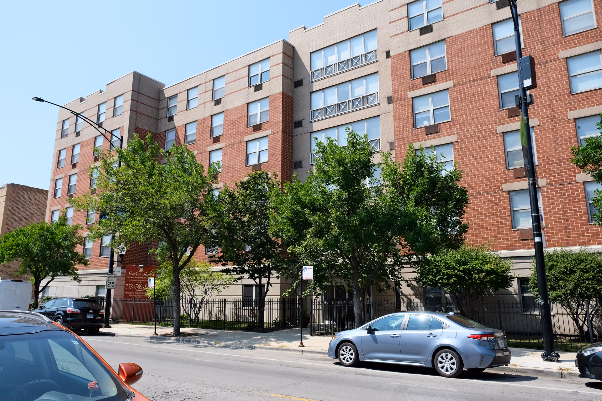 Exterior view of a multi-story brick and concrete senior living facility building with several windows, trees lining the sidewalk, and parked cars along the street in front.