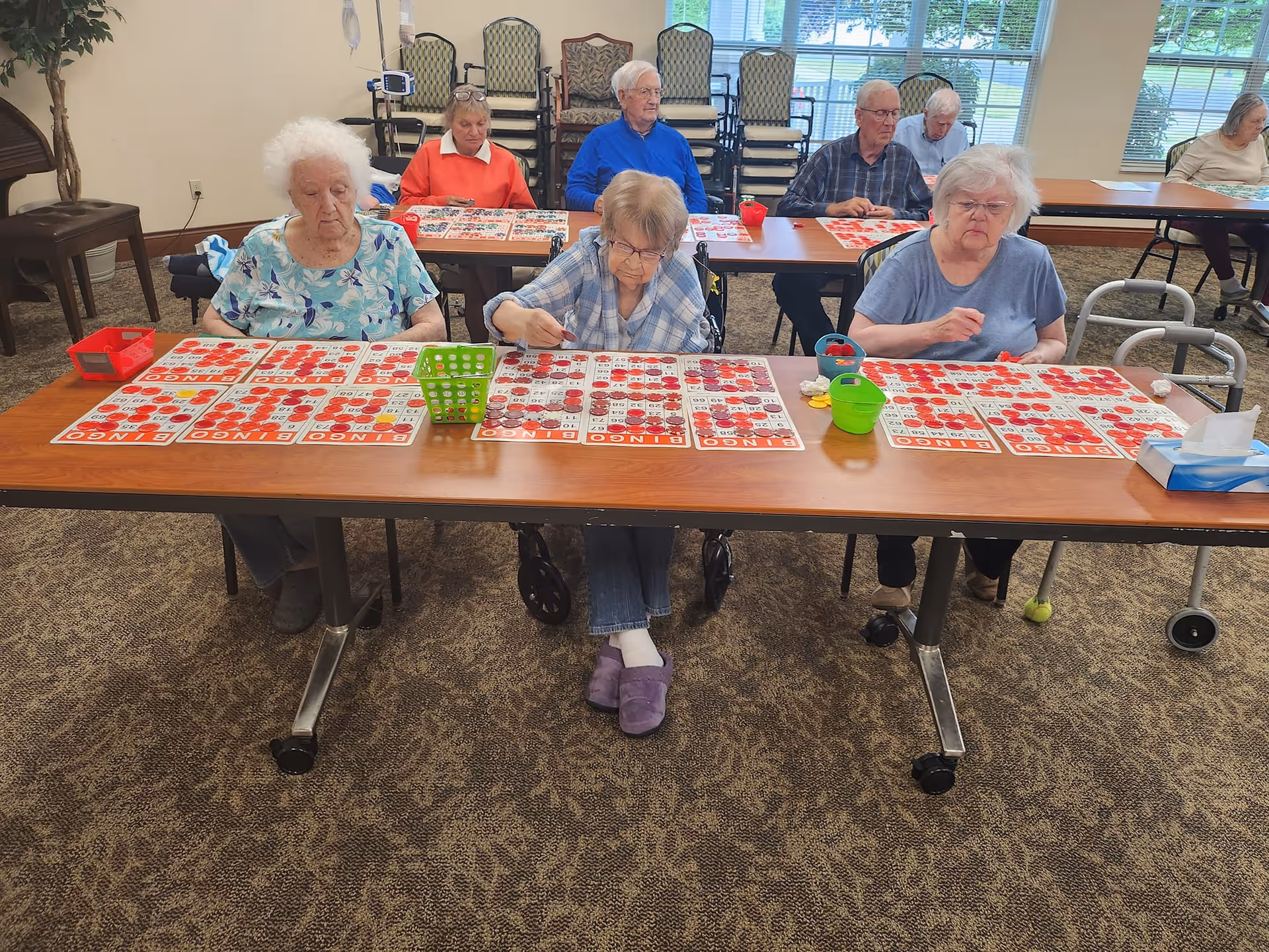 A group of elderly residents sitting at tables in a community room playing bingo. Several bingo cards with red markers are spread out in front of them. The room has carpeted floors, large windows, and stacked chairs in the background.