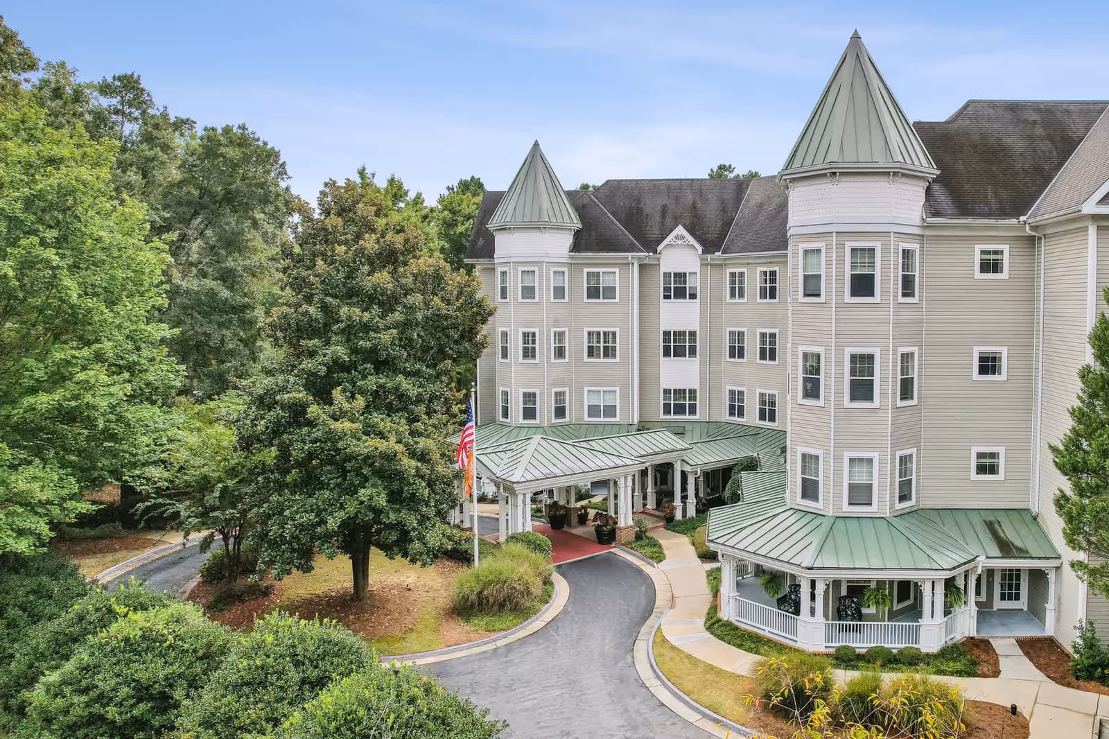 Exterior view of a senior living facility named Sunrise of Decatur, featuring a large multi-story building with light-colored siding, green metal roofs, and two turret-like structures. The entrance has a covered driveway with a red carpet, surrounded by well-maintained landscaping and trees under a clear sky.