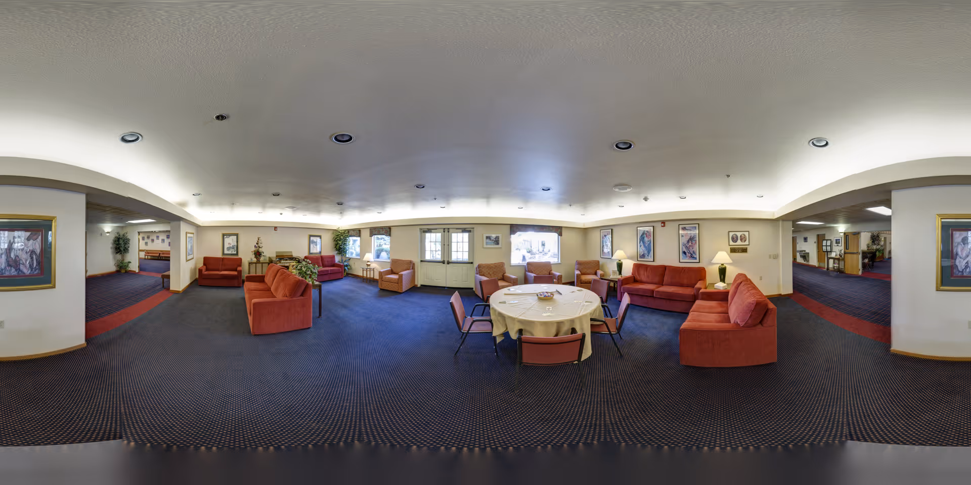 A spacious common area in a senior living facility with multiple red upholstered sofas and armchairs arranged around a central round table covered with a white tablecloth. The room has blue carpet with a red stripe, white walls adorned with framed artwork, and several lamps providing warm lighting. There are windows and a door letting in natural light, and the ceiling has recessed lighting.