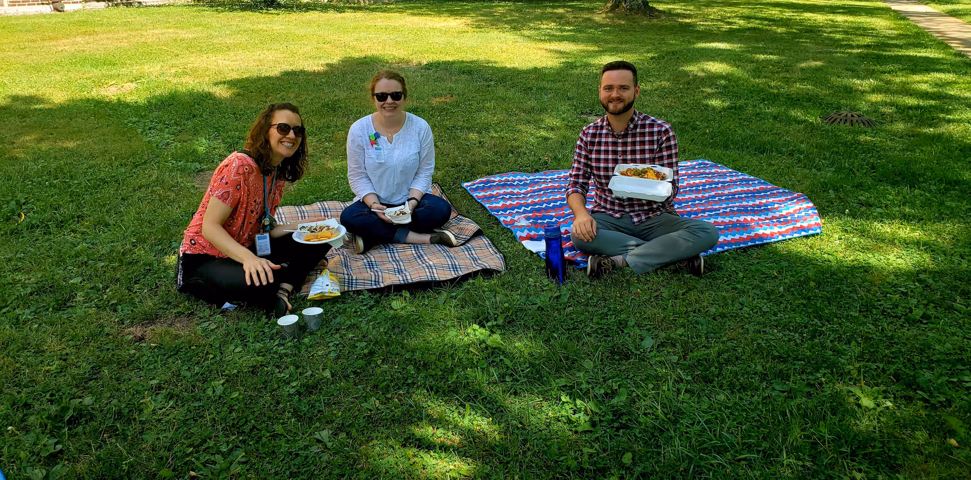 Three people sitting on blankets on a grassy lawn having a picnic. Two women and one man are smiling at the camera while holding plates of food. The setting is outdoors with green grass and some shaded areas.