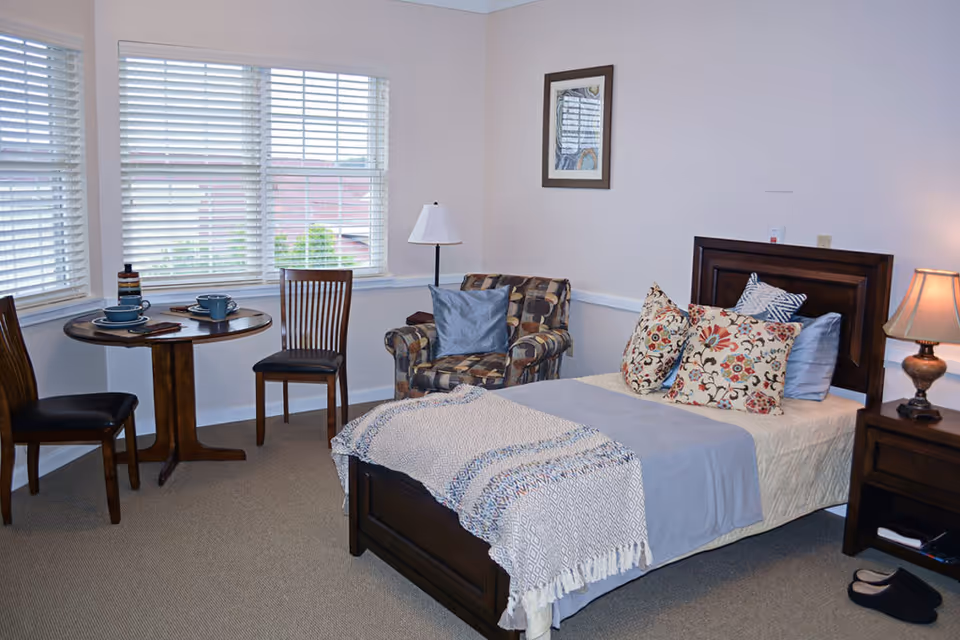A cozy bedroom in a senior living facility with a single bed adorned with multiple decorative pillows and a light blue blanket. Next to the bed is a wooden nightstand with a lamp and slippers on the floor. There is a patterned armchair with a blue cushion, a small round wooden table set with two place settings, and two wooden chairs near a window with white blinds. A framed picture hangs on the wall above the armchair.