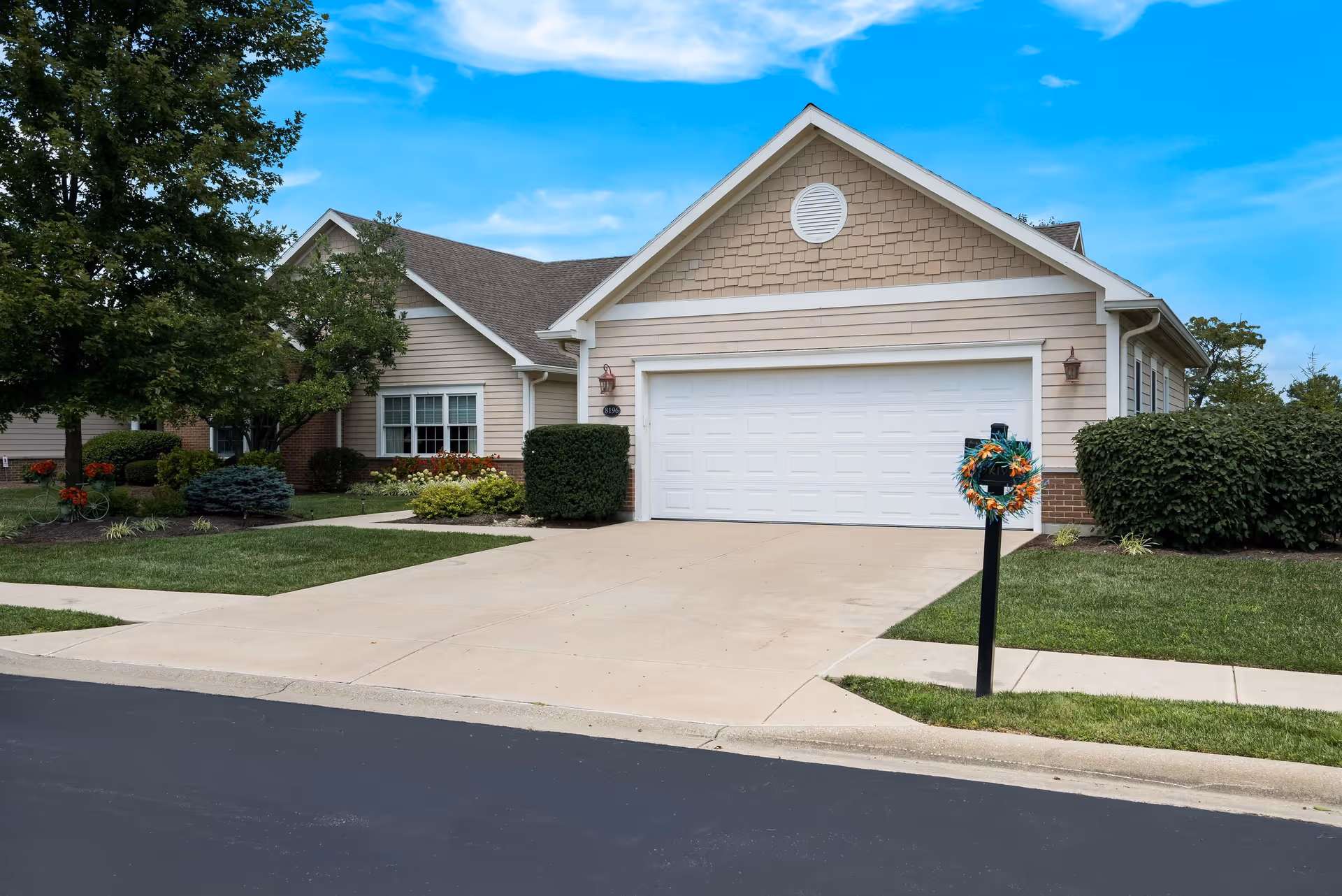 Exterior view of a single-story residential building with beige siding and a white garage door. The driveway is clean and leads to the garage. There is a mailbox with a decorative wreath on a post near the driveway. The lawn is well-maintained with green grass, bushes, and trees under a partly cloudy blue sky.