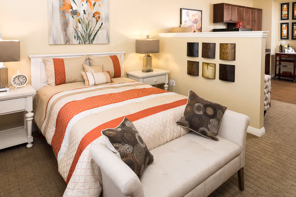 A cozy bedroom featuring a neatly made bed with a beige and orange striped quilt, two matching pillows, and a cushioned bench with patterned pillows at the foot of the bed. The room includes two white nightstands with lamps, a wall painting of flowers above the bed, and a partial view of a kitchen area separated by a half wall with decorative square wall art.
