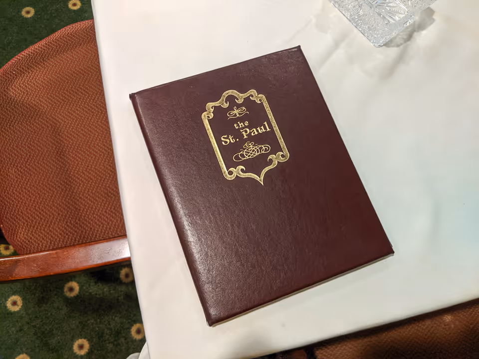 A maroon leather-bound menu or book with gold embossed text and decorative border that reads 'the St. Paul' placed on a white tablecloth. Part of a reddish-brown chair and a green carpet with a floral pattern are visible beside the table.