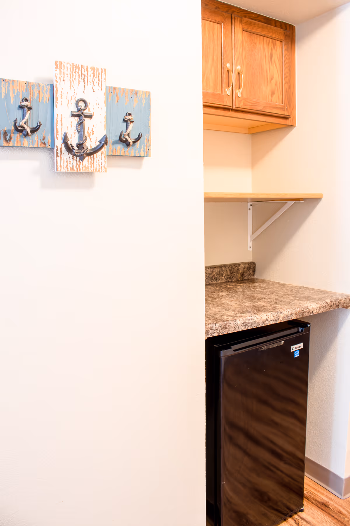 Small kitchenette nook with a mini fridge beneath a laminate countertop, wooden cabinets, shelf, and anchor-themed wall art.