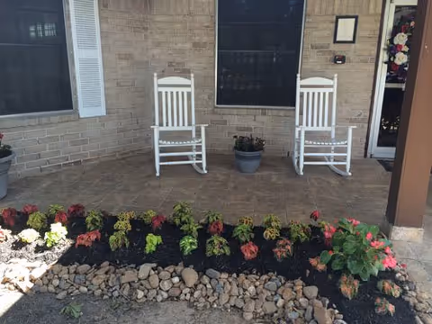 Front porch with two white rocking chairs, potted plants, and a flowerbed edged with rocks in front of a brick wall and entrance.