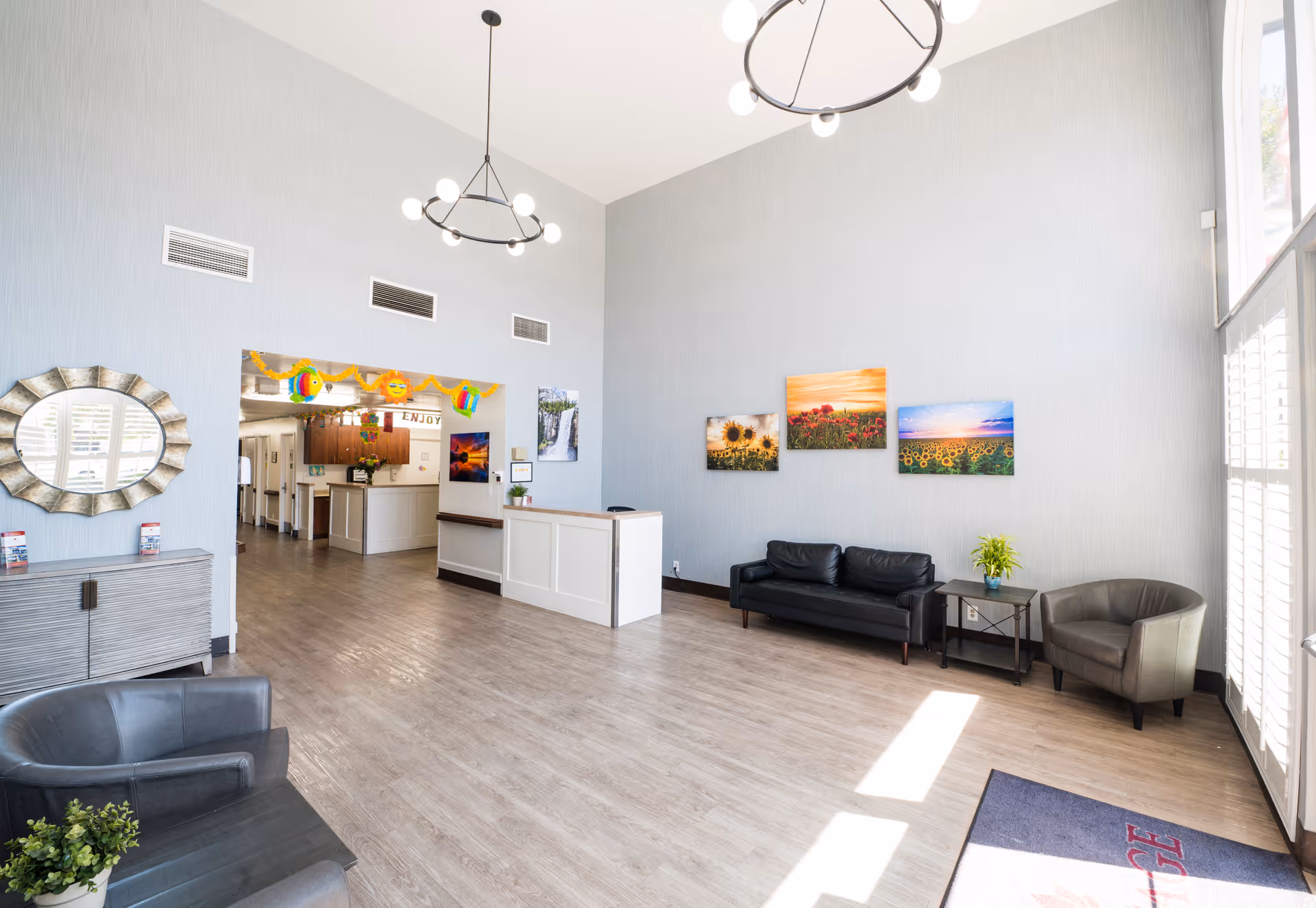 A bright and spacious senior living facility lobby with light gray walls and wood flooring. The room features modern black leather seating including a sofa and two armchairs, a small side table with a plant, and colorful nature-themed artwork on the walls. There is a reception desk in the background with a hallway leading further into the facility. Large windows with white shutters allow natural light to fill the space. Two modern circular chandeliers hang from the high ceiling.