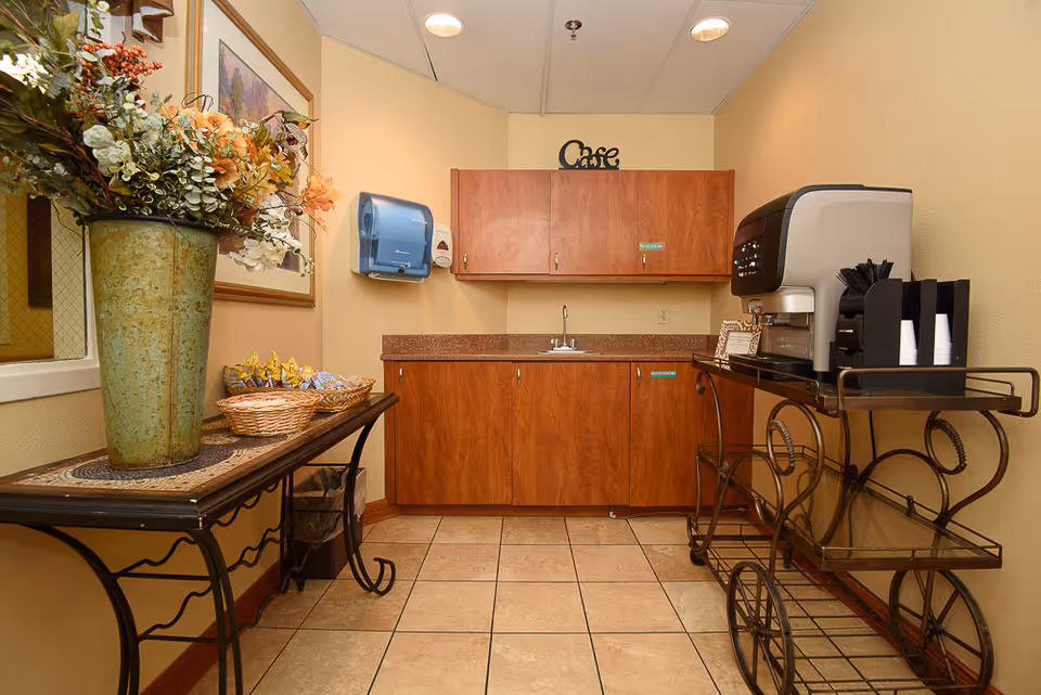 Small kitchenette area with wooden cabinets, a countertop with a sink, a wall-mounted paper towel dispenser, and a coffee machine on a metal cart. A decorative table with a large vase of flowers and baskets of snacks is on the left side.