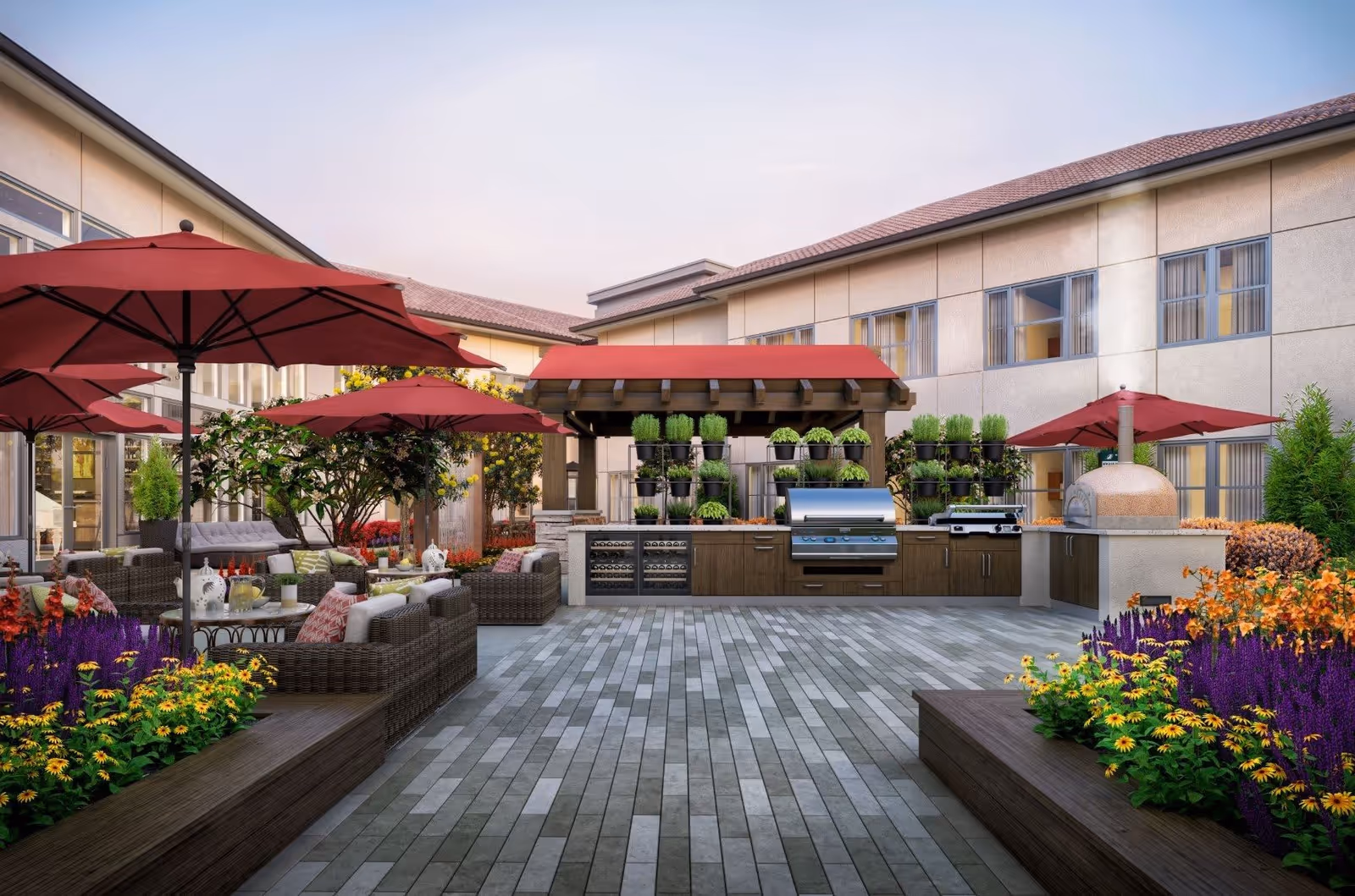 Outdoor patio area at Varenita Simi Valley featuring multiple red umbrellas shading wicker seating arrangements with cushions. There are raised garden beds with colorful flowers and a built-in outdoor kitchen with a grill, pizza oven, and herb planters under a pergola with a red canopy.