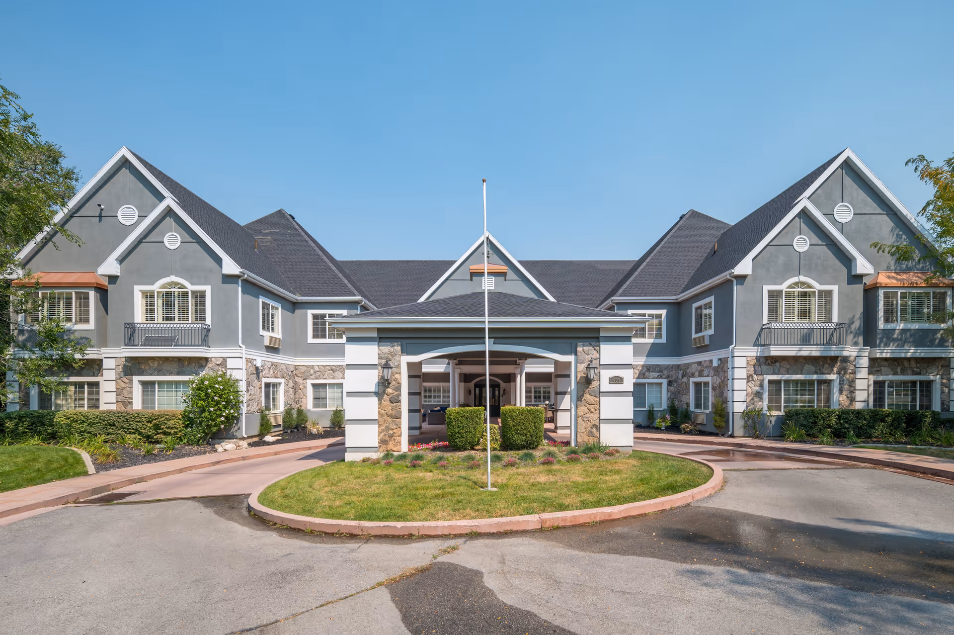 Front exterior view of a senior living facility building with a driveway, landscaped greenery, and a covered entrance under a clear blue sky.