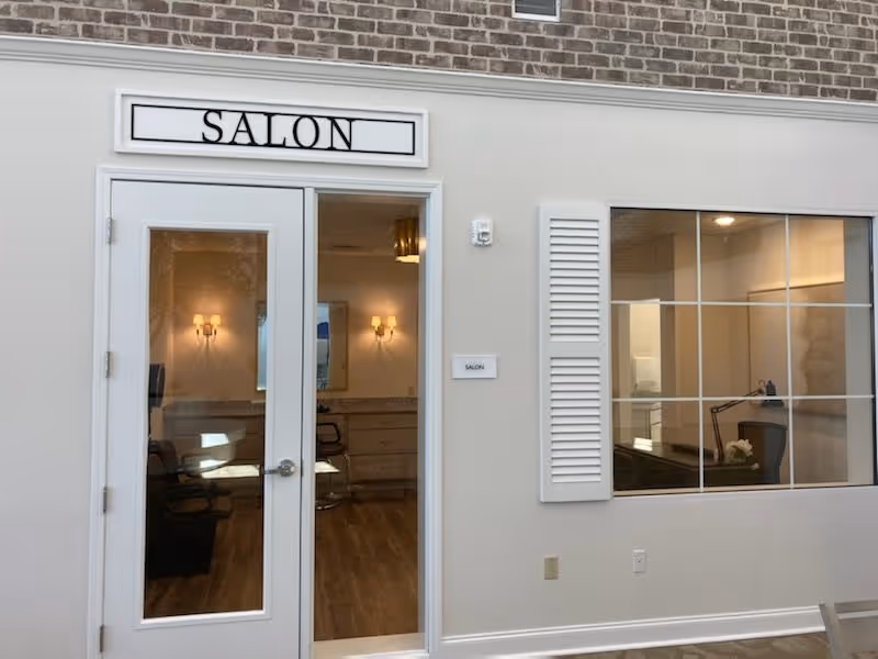 Entrance to a salon room in a senior living facility with double glass doors and a sign above reading 'SALON'. Inside, salon chairs and mirrors with wall-mounted lights are visible. The wall next to the door has a large window with white shutters, showing an office area with a desk and lamp.